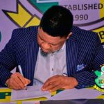 Coach Daniel Ogunmodede in a pinstripe suit signs his contract at a table during Kano Pillars Unveiling event with logos visible in the background and a crest on the table  trybalnews