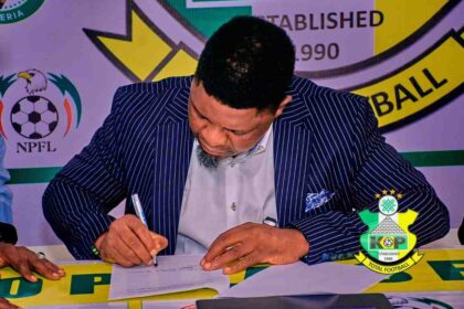Coach Daniel Ogunmodede in a pinstripe suit signs his contract at a table during Kano Pillars Unveiling event, with logos visible in the background and a crest on the table.