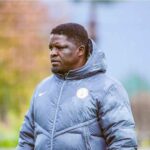 Daniel Ogunmodede assistant coach of the Super Eagles of Nigeria stands on a football training pitch wearing a grey Nike puffer jacket with the Nigeria Football Federation crest and black gloves He has a focused expression and is looking to his left The background is blurred with a training field and trees He has been linked with the Kano Pillars coaching job  trybalnews