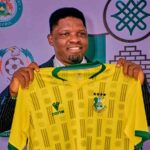 New Kano Pillars Technical Adviser Daniel Ogunmodede holding up a yellow and green Kano Pillars jersey at his official press unveiling He wears a navy pinstripe suit and smiles at the camera  trybalnews