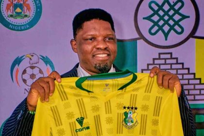 New Kano Pillars Technical Adviser Daniel Ogunmodede holding up a yellow and green Kano Pillars jersey at his official press unveiling. He wears a navy pinstripe suit and smiles at the camera.