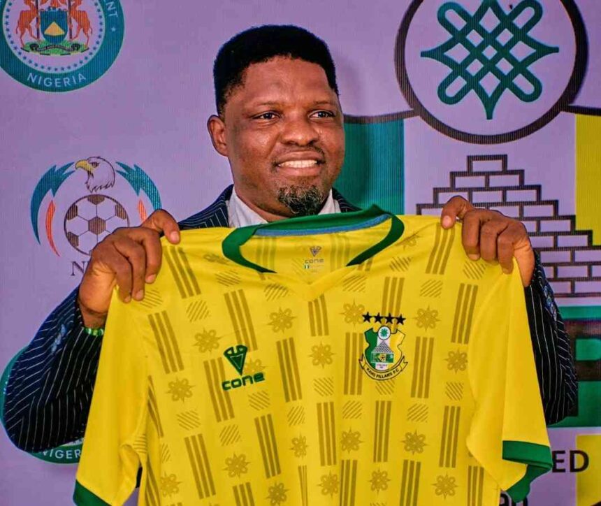 New Kano Pillars Technical Adviser Daniel Ogunmodede holding up a yellow and green Kano Pillars jersey at his official press unveiling He wears a navy pinstripe suit and smiles at the camera  trybalnews