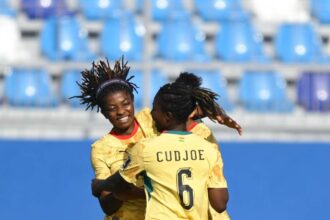 Two soccer players in yellow jerseys hug and celebrate on the field with blue stadium seating behind them; one jersey shows the name CUDJOE and number 6.
