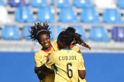 Two soccer players in yellow jerseys hug and celebrate on the field with blue stadium seating behind them; one jersey shows the name CUDJOE and number 6.