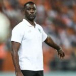 Emerse Fae in a white polo shirt and black shorts standing on a soccer field with an orange clad crowd in the background  trybalnews