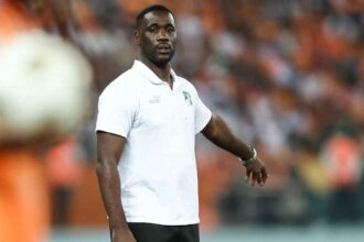 Emerse Fae in a white polo shirt and black shorts standing on a soccer field with an orange-clad crowd in the background.