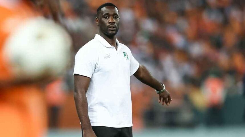 Emerse Fae in a white polo shirt and black shorts standing on a soccer field with an orange clad crowd in the background  trybalnews