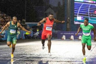 Cameroon's Emmanuel Eseme in red sprints to gold in the Men's 100m final at Scotstoun Stadium during the 2026 Commonwealth Games in Glasgow, Scotland on July 28, 2026, ahead of South Africa's Akani Simbine in green and Nigeria's Kayinsola Ajayi in green and white, on a wet rain-soaked track.