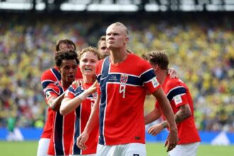 Norway striker Erling Haaland wearing jersey number 9 celebrates with teammates after scoring his team's first goal. The players are grouped together on the pitch at New York New Jersey Stadium during the FIFA World Cup 2026 Round of 16 match between Brazil and Norway on July 5, 2026. Stadium crowd and green turf are visible in the background.