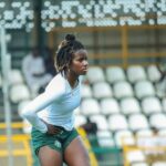 Female athlete standing with hands on hips on a track field wearing a white long sleeve top and green shorts blurred stadium seats in the background  trybalnews