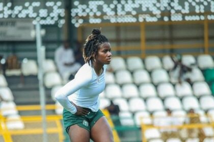 Female athlete standing with hands on hips on a track field, wearing a white long-sleeve top and green shorts, blurred stadium seats in the background.