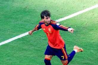 Ferran Torres of Spain, wearing a red jersey with number 7, celebrates on the green pitch with arms outstretched and one leg lifted after scoring Spain's first goal in the FIFA World Cup 2026 Final against Argentina at MetLife Stadium in East Rutherford, New Jersey.