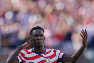 USA forward Folarin Balogun, wearing the #20 red and white striped home kit, celebrates with his finger to his ear and one arm raised after scoring his team's first goal during the FIFA World Cup 2026 Round of 32 match against Bosnia and Herzegovina at San Francisco Bay Area Stadium in Santa Clara, California, on July 1, 2026.