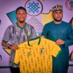 Two men stand side by side holding a yellow Nigeria national team jersey with green trim and crest against a backdrop of football association logos  trybalnews