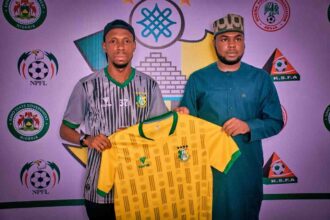 Two men stand side by side holding a yellow Nigeria national team jersey with green trim and crest, against a backdrop of football association logos.