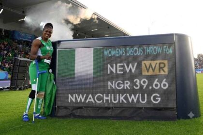 Gold medalist Goodness Nwachukwu of Team Nigeria in green and white kit stands next to a Longines scoreboard at Scotstoun Stadium, holding a Nigerian flag and crutches, after setting a new world record of 39.66m in the Women's Discus Throw F42-44/F61-64 Final on Day 6 of the Glasgow 2026 Commonwealth Games, July 29, 2026.