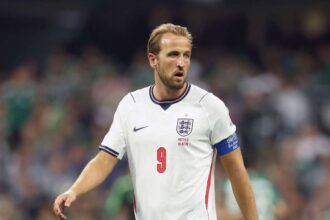 England captain Harry Kane wearing jersey number 9 and a white England kit with navy shorts walks on the pitch during play. He wears a blue captain's armband and looks ahead with a focused expression during the FIFA World Cup 2026 Round of 16 match between Mexico and England at Mexico City Stadium on July 5, 2026. Blurred crowd and players in green are visible in the background.