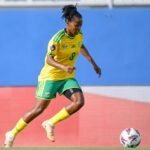 Female soccer player in Brazil kit yellow jersey green shorts dribbles toward the ball on a soccer field during a match with a blue backdrop behind  trybalnews
