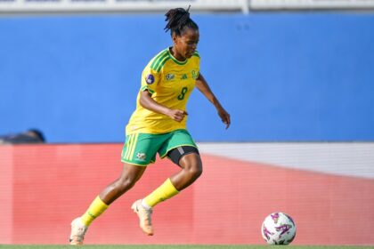 Female soccer player in Brazil kit (yellow jersey, green shorts) dribbles toward the ball on a soccer field during a match, with a blue backdrop behind.
