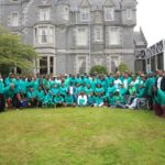 Large group of people wearing teal shirts posing for a group photo on a grassy lawn in front of a gray castle like building with towers  trybalnews