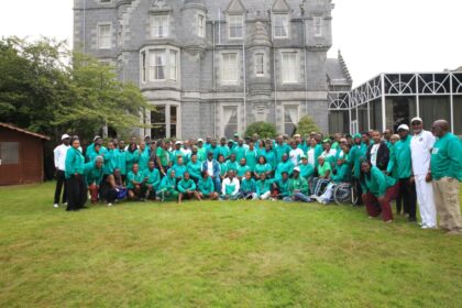 Large group of people wearing teal shirts posing for a group photo on a grassy lawn in front of a gray castle-like building with towers.