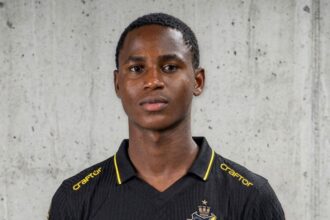 Portrait of a young man in a black AIK football jersey with yellow trim, standing against a concrete wall. The kit features the AIK crest and the sponsor 'truecaller' in yellow.