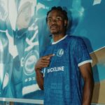 Young man in a blue patterned soccer jersey with BACKLINE logo pointing to himself stands against a teal mural background  trybalnews