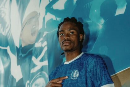 Young man in a blue patterned soccer jersey with 'BACKLINE' logo, pointing to himself, stands against a teal mural background.