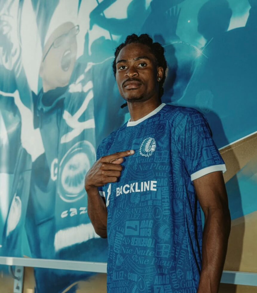 Young man in a blue patterned soccer jersey with BACKLINE logo pointing to himself stands against a teal mural background  trybalnews