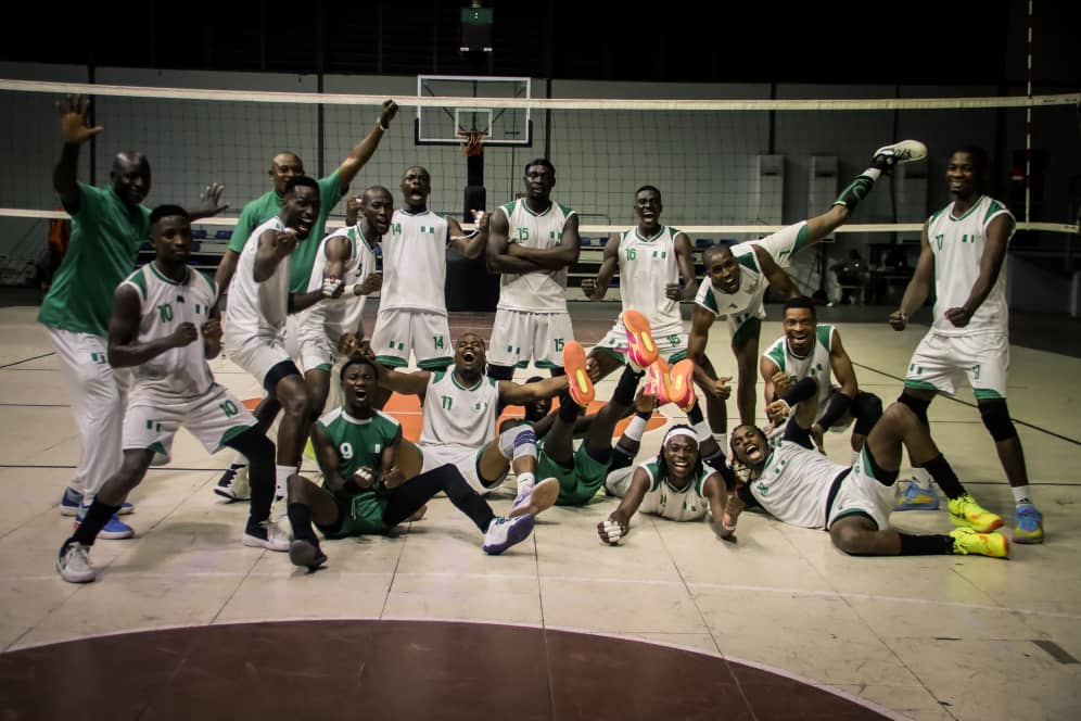 IMG20260717WA0004 Trybal News A menaposs volleyball team in white and green uniforms celebrates on an indoor court with players standing kneeling and some kicking their legs in front of a net trybalnews