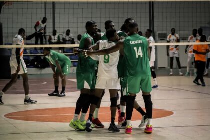Indoor team sport scene with players in green and white uniforms gathering in a huddle near the net, celebrating or encouraging each other on a court.
