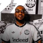 Portrait of a man in a white Eintracht Frankfurt jersey with round glasses posed in front of a wall of black and white posters  trybalnews