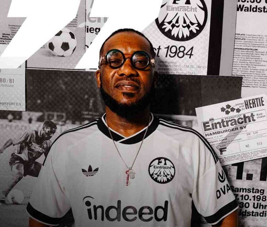 Portrait of a man in a white Eintracht Frankfurt jersey with round glasses posed in front of a wall of black and white posters  trybalnews