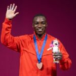 Medalist in orange waving on stage wearing a blue ribbon medal and holding a pink unicorn plush  trybalnews