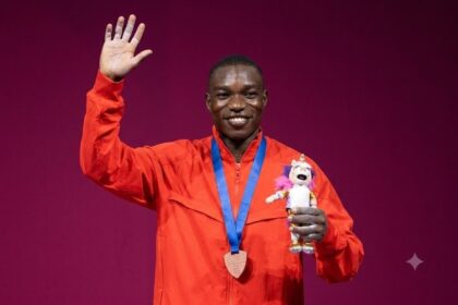Medalist in orange waving on stage, wearing a blue ribbon medal and holding a pink unicorn plush.