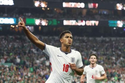 England midfielder Jude Bellingham wearing a white jersey number 10 and navy shorts points to the sky with his right arm as he celebrates. He has just scored England's first goal during the FIFA World Cup 2026 Round of 16 match between Mexico and England at Mexico City Stadium on July 5, 2026. England teammate Declan Rice wearing number 4 runs behind him, with a packed stadium and "MEXICO CITY" on the scoreboard in the background.