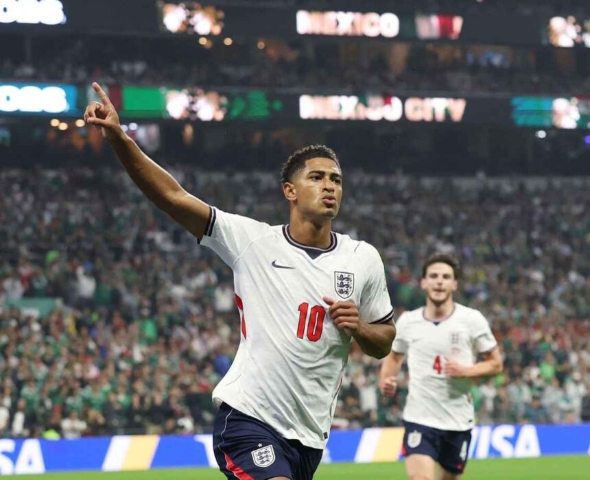 England midfielder Jude Bellingham wearing a white jersey number 10 and navy shorts points to the sky with his right arm as he celebrates He has just scored Englands first goal during the FIFA World Cup 2026 Round of 16 match between Mexico and England at Mexico City Stadium on July 5 2026 England teammate Declan Rice wearing number 4 runs behind him with a packed stadium and MEXICO CITY on the scoreboard in the background  trybalnews
