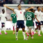 England midfielder Jude Bellingham wearing a white jersey number 10 and navy shorts stands on the pitch with both arms outstretched celebrating after scoring Englands second goal Behind him England and Mexico players are visible during the FIFA World Cup Round of 16 match at Mexico City Stadium on Sunday July 5 2026 A Mexico player wearing green jersey number 25 R Alvarado walks away in the foreground  trybalnews