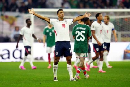 England midfielder Jude Bellingham wearing a white jersey number 10 and navy shorts stands on the pitch with both arms outstretched, celebrating after scoring England’s second goal. Behind him, England and Mexico players are visible during the FIFA World Cup Round of 16 match at Mexico City Stadium on Sunday, July 5, 2026. A Mexico player wearing green jersey number 25, R. Alvarado, walks away in the foreground.
