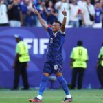 France forward Kylian Mbappe wearing blue jersey number 10 celebrates with both arms raised and index fingers pointing up after scoring a penalty He stands on the green pitch at Philadelphia Stadium during the FIFA World Cup 2026 Round of 16 match between France and Paraguay on July 4 2026 Stadium staff in yellow vests and fans are blurred in the background with a large blue FIFA advertising board behind him  trybalnews