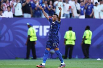 France forward Kylian Mbappe wearing blue jersey number 10 celebrates with both arms raised and index fingers pointing up after scoring a penalty. He stands on the green pitch at Philadelphia Stadium during the FIFA World Cup 2026 Round of 16 match between France and Paraguay on July 4, 2026. Stadium staff in yellow vests and fans are blurred in the background, with a large blue FIFA advertising board behind him.