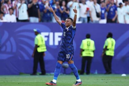 France forward Kylian Mbappe wearing blue jersey number 10 celebrates with both arms raised and index fingers pointing up after scoring a penalty. He stands on the green pitch at Philadelphia Stadium during the FIFA World Cup 2026 Round of 16 match between France and Paraguay on July 4, 2026. Stadium staff in yellow vests and fans are blurred in the background, with a large blue FIFA advertising board behind him.