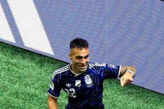 Soccer player wearing blue kit with number 22 celebrating on green artificial turf, fists raised, near side line; teammate in a yellow vest watches.