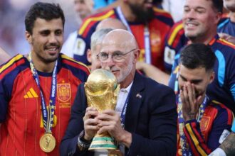 Spain head coach Luis de la Fuente, wearing a navy suit and glasses, holds and kisses the golden FIFA World Cup trophy while celebrating with smiling Spain players in red jerseys after the FIFA World Cup 2026 Final at New York New Jersey Stadium in East Rutherford, New Jersey.