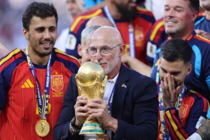 Spain head coach Luis de la Fuente, wearing a navy suit and glasses, holds and kisses the golden FIFA World Cup trophy while celebrating with smiling Spain players in red jerseys after the FIFA World Cup 2026 Final at New York New Jersey Stadium in East Rutherford, New Jersey.