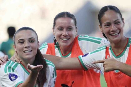 Three female soccer players celebrate together on the field, arms around each other and smiling, wearing white and green jerseys with orange bibs.