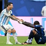 Lionel Messi of Argentina wearing the light blue and white striped jersey smiles while helping up an opponent in a dark blue jersey with LOPEZ and number 11 on the back The opponent is sitting on the grass holding Messis hands after a match A football sits nearby on the pitch with advertising boards and stadium staff in the background  trybalnews