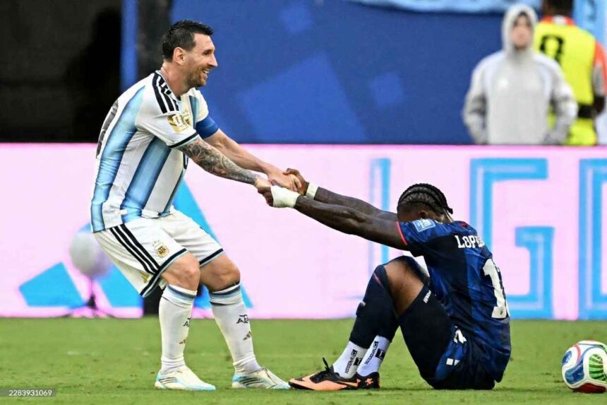 Lionel Messi of Argentina wearing the light blue and white striped jersey smiles while helping up an opponent in a dark blue jersey with LOPEZ and number 11 on the back The opponent is sitting on the grass holding Messis hands after a match A football sits nearby on the pitch with advertising boards and stadium staff in the background  trybalnews