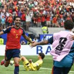 Spain footballer wearing red jersey 6 celebrates with arms outstretched as teammates run toward him crowd cheers in the stands behind  trybalnews