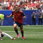 Spains 21 celebrates a goal in red kit with pink boots running on the field as an England player sits on the grass nearby  trybalnews
